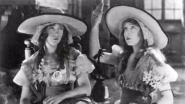 Henriette (Lillian Gish) and Louise (Dorothy Gish) sit together indoors beneath broad hats, holding flowers