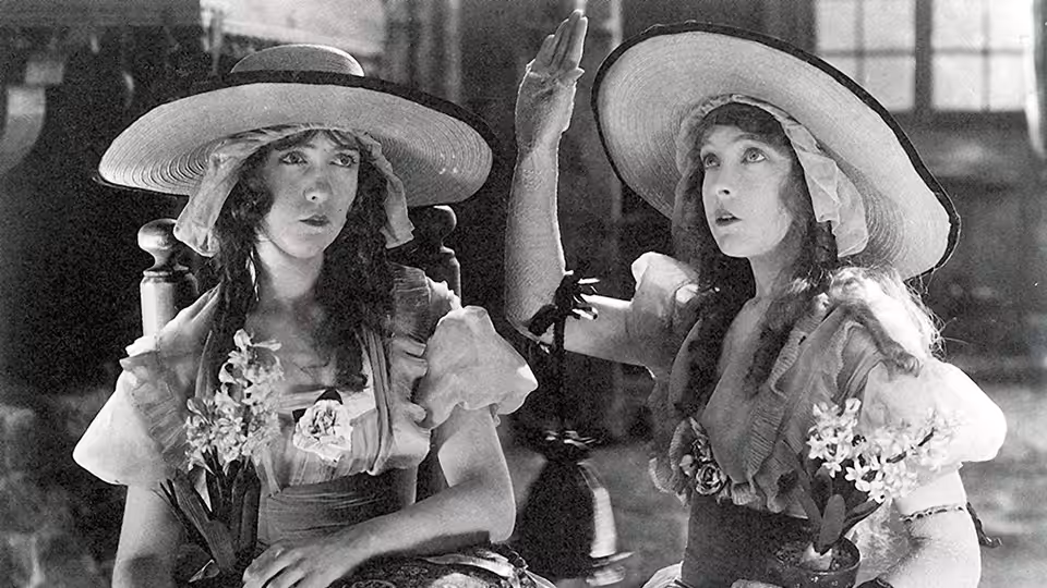 Henriette (Lillian Gish) and Louise (Dorothy Gish) sit together indoors beneath broad hats, holding flowers