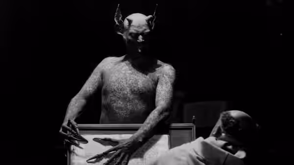 The horned Devil stands behind an open chest as a figure lies inside