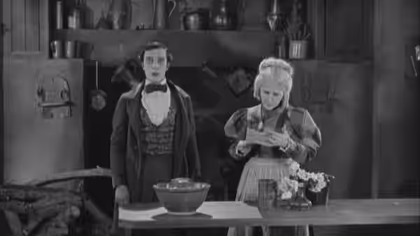 William McKay (Buster Keaton) stands beside a woman preparing food at a kitchen table