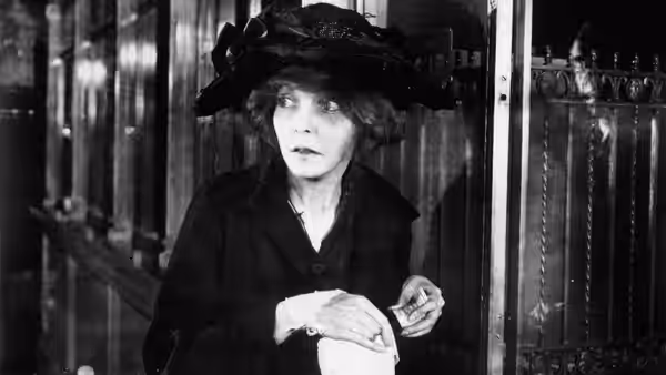 A woman in a dark hat holds a small object beside a row of shop windows