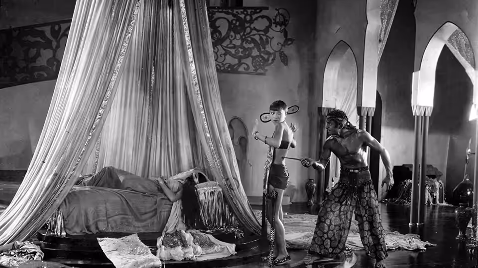 A turbaned man stands beside a curtained bed where a woman reclines
