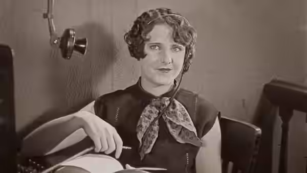 A curly-haired person in a patterned scarf sits at a desk holding a paper