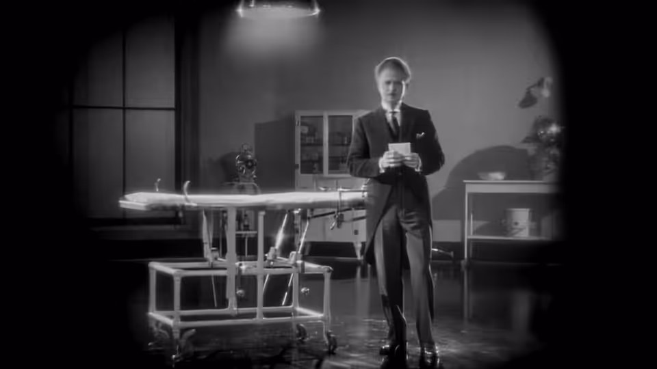 A suited man stands among tables and equipment in a dim room