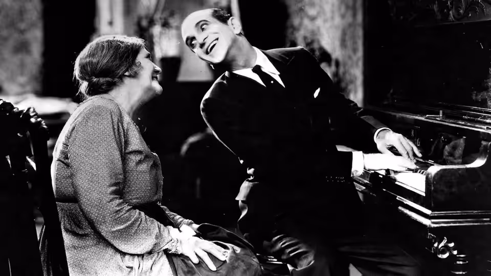Jakie Rabinowitz (Al Jolson) leans over a seated woman beside a piano