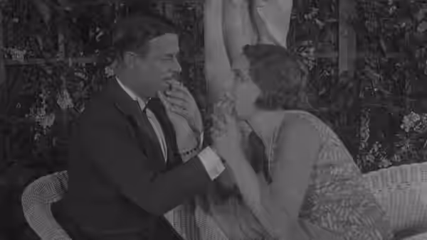 A suited man and woman sit close, holding hands near their mouths