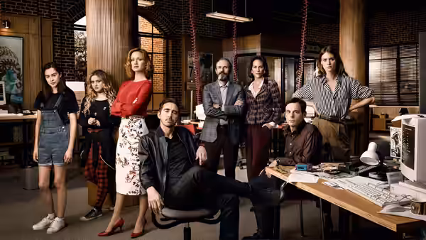 Lee Pace seated in foreground with six colleagues standing in an office