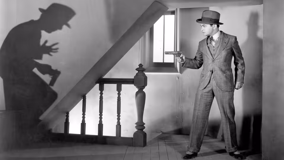 A suited man stands beside a staircase, aiming a pistol toward a shadow on the wall