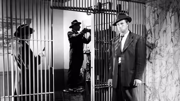 A suited man stands by barred doors as an officer watches