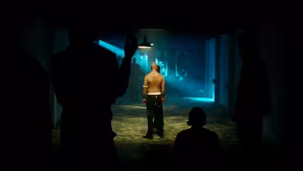 A shirtless man stands in a blue-lit room as shadowed figures watch