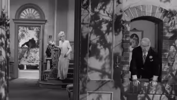 A woman stands by the staircase as an older man enters through a doorway