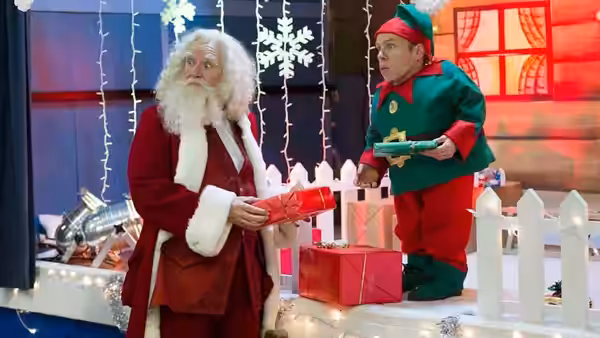 Santa Claus (Jim Broadbent) exchanges a gift with an elf beside a festive display