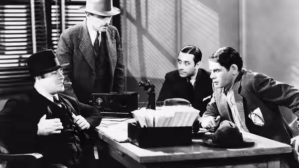 Tony Camonte (Paul Muni) leans across a desk toward two men in an office