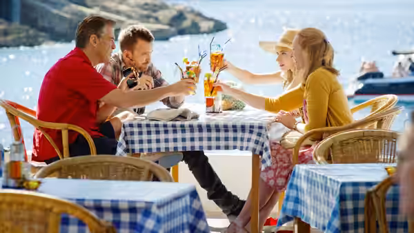 Four people sit around a checkered café table, sharing drinks beside the water