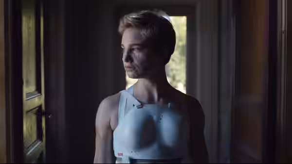 A short-haired person wearing a white chest brace stands in a dim doorway