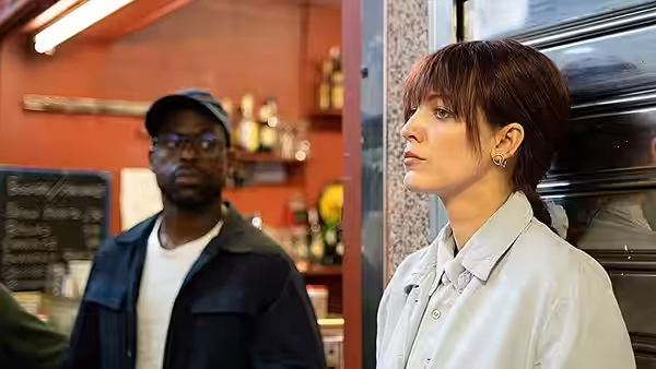 A woman in a white coat stands beside a man in a cap inside a shop