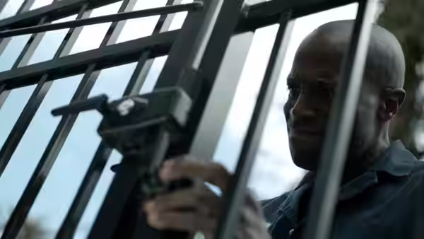 A bald man looks through a barred window, one hand raised beside the frame