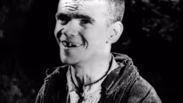 A smiling man in a jacket faces the camera against a dark background