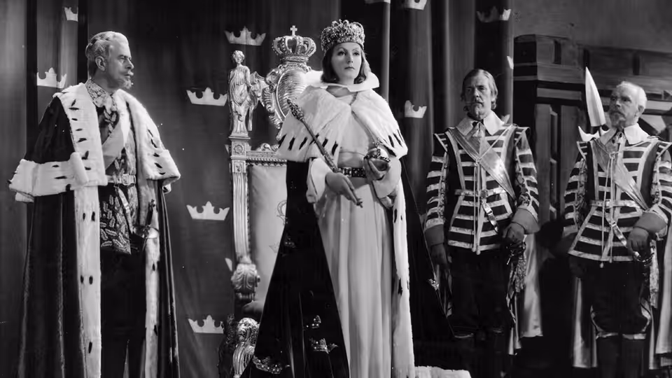 Christina (Greta Garbo) stands crowned among courtiers and uniformed guards in the palace