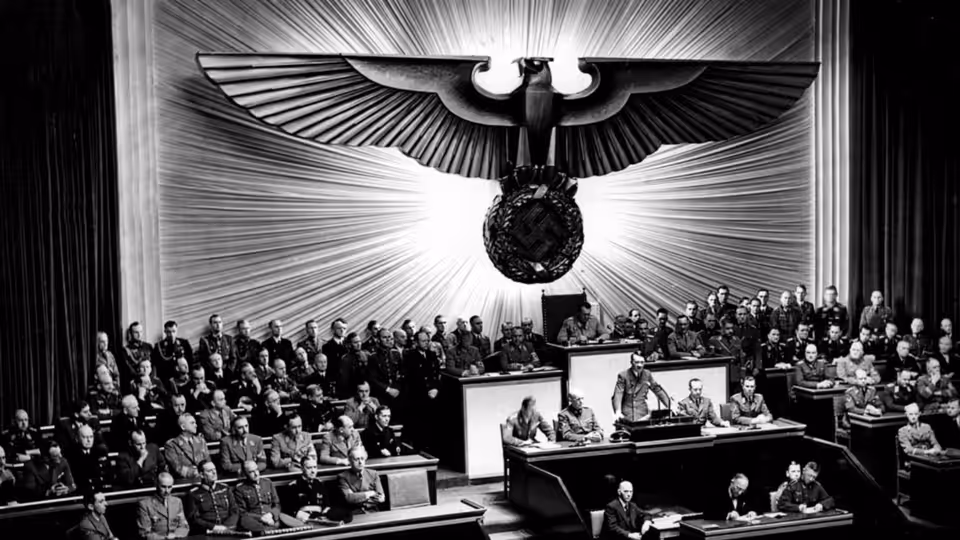 Officials sit beneath a monumental eagle emblem in a crowded assembly hall