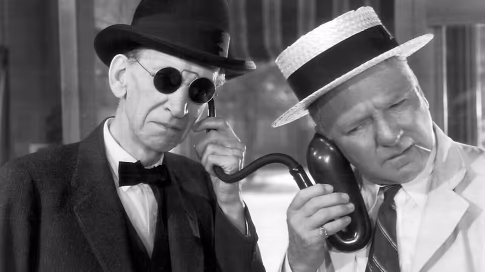 A man in a light suit listens to a telephone while another man holds a pipe beside him