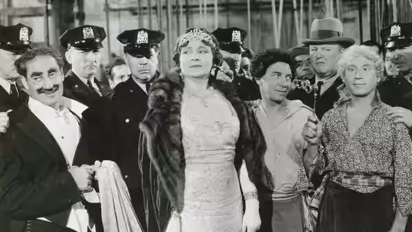 Groucho Marx stands beside a formally dressed woman as uniformed officers gather around them