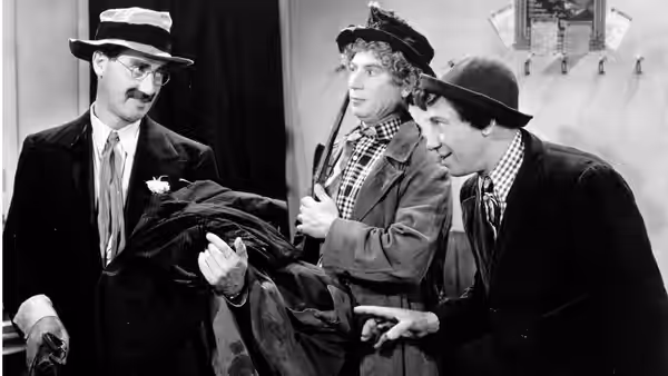 Chico Marx and Harpo Marx carry a bundled passenger past a mustached uniformed man
