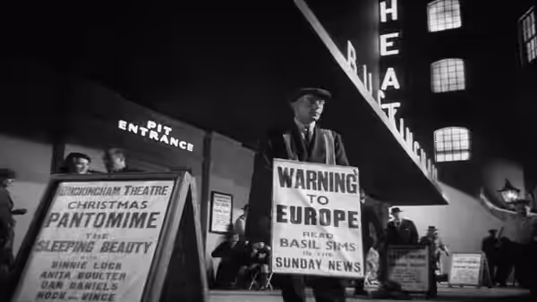 A man stands among street-side news placards and theatre advertisements
