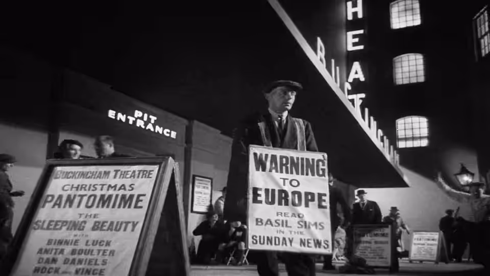 A man stands among street-side news placards and theatre advertisements