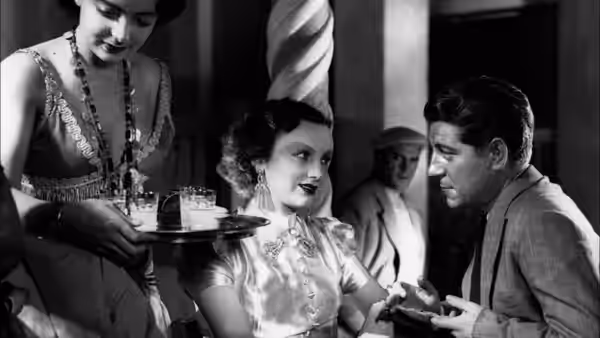 Pépé le Moko (Jean Gabin) speaks with a seated woman as a server offers refreshments