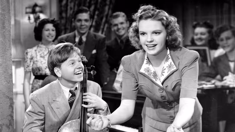 Mickey Moran (Mickey Rooney) sits with a cello as Patsy Barton (Judy Garland) leans forward beside him