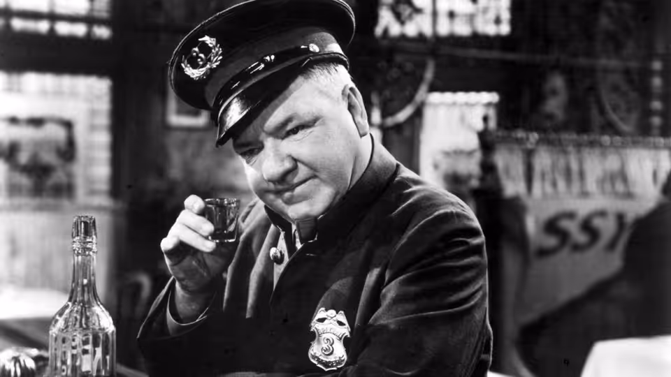 Egbert Sousé (W.C. Fields) in a police uniform holds a glass beside a bar counter
