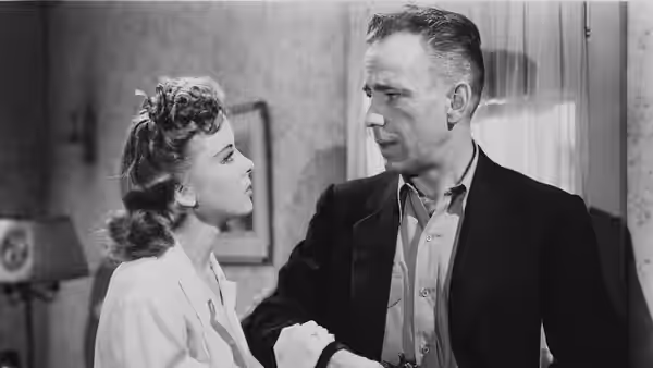 Roy Earle (Humphrey Bogart) looks at a young woman beside him indoors