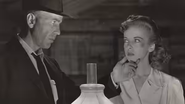 A man in a fedora and a young woman stand beside a lamp