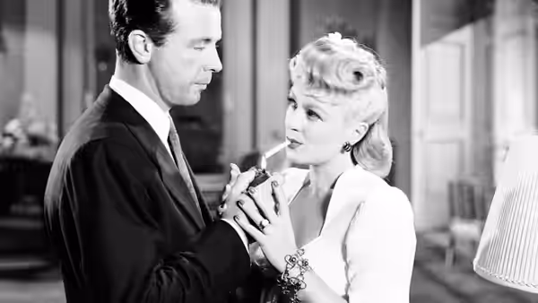 A suited man and a blonde woman stand close, holding hands beside a lamp