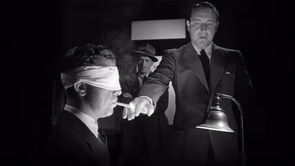 A blindfolded man sits beside a lamp as a suited man points toward him