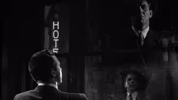 A seated man looks toward a hotel sign and a suited man reflected in glass