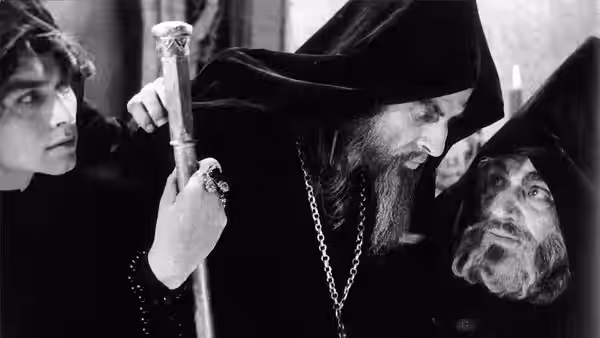 A hooded, bearded man holds a staff between two men