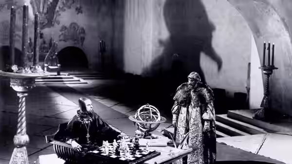 A seated man faces another man across a chessboard in an ornate hall