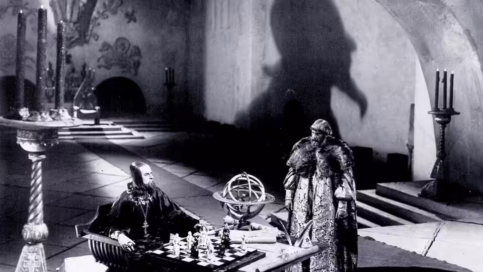 A seated man faces another man across a chessboard in an ornate hall