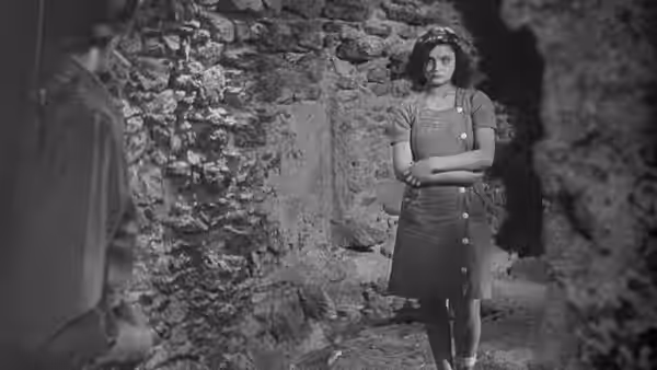 A young woman stands with folded arms in a stone doorway as a man faces her