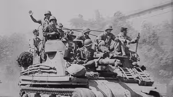 Soldiers ride an armored vehicle, one waving as it moves through the countryside
