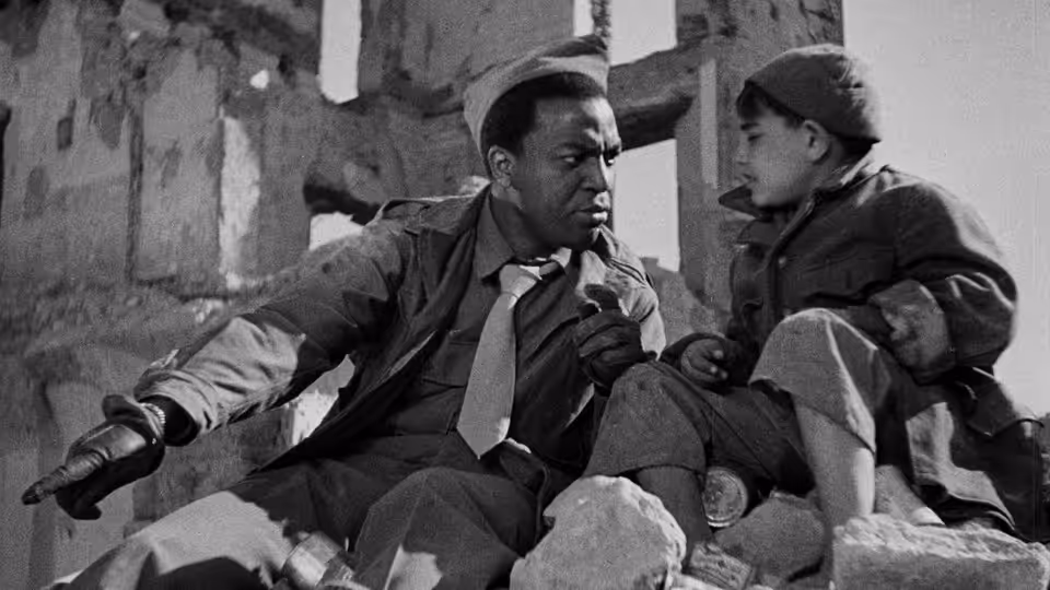 An American soldier and a young boy sit among rubble in a ruined city