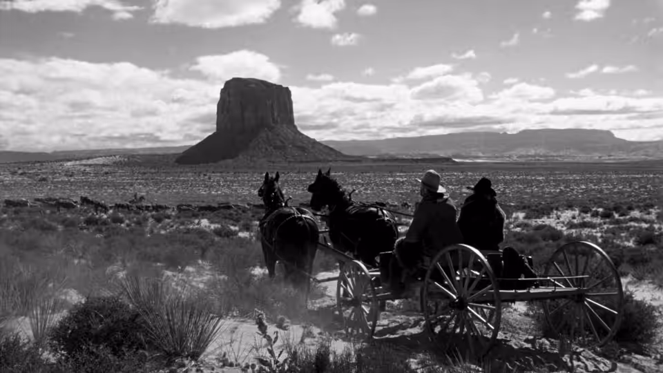 A covered wagon and riders cross a wide, open desert plain
