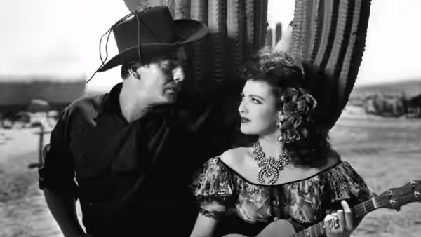 A cowboy stands beside a woman holding a guitar near a curtained window