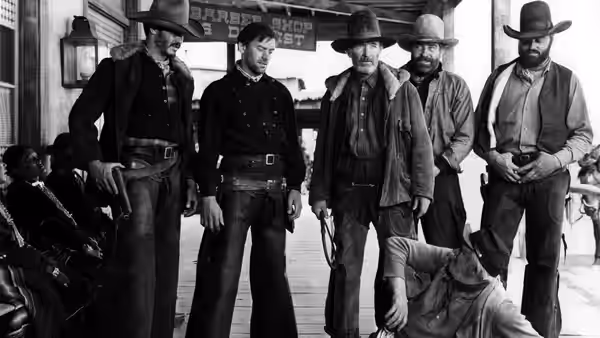 Four armed cowboys stand outside a storefront as two men lie on the ground
