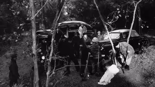 Several men gather beside a parked car as another figure crouches near a body on the ground