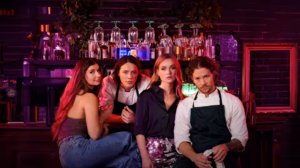 Three people gather behind a bar counter as a man in an apron holds a cloth