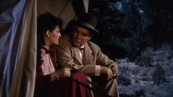 Calamity Jane sits beside Peter Potter in a hat and suit, looking toward him at night
