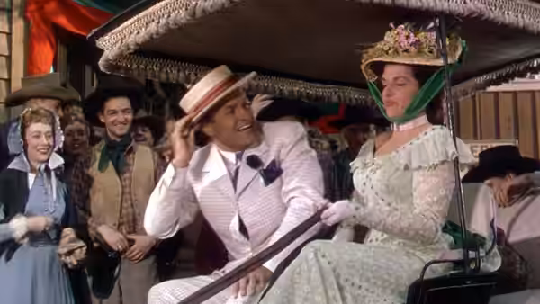Peter Potter tips his hat beside Calamity Jane in a carriage as onlookers gather